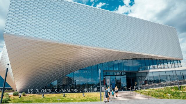 The U.S. Olympic & Paralympic Museum with a modern, geometric exterior design, visitors approaching the entrance on a sunny day with blue skies.